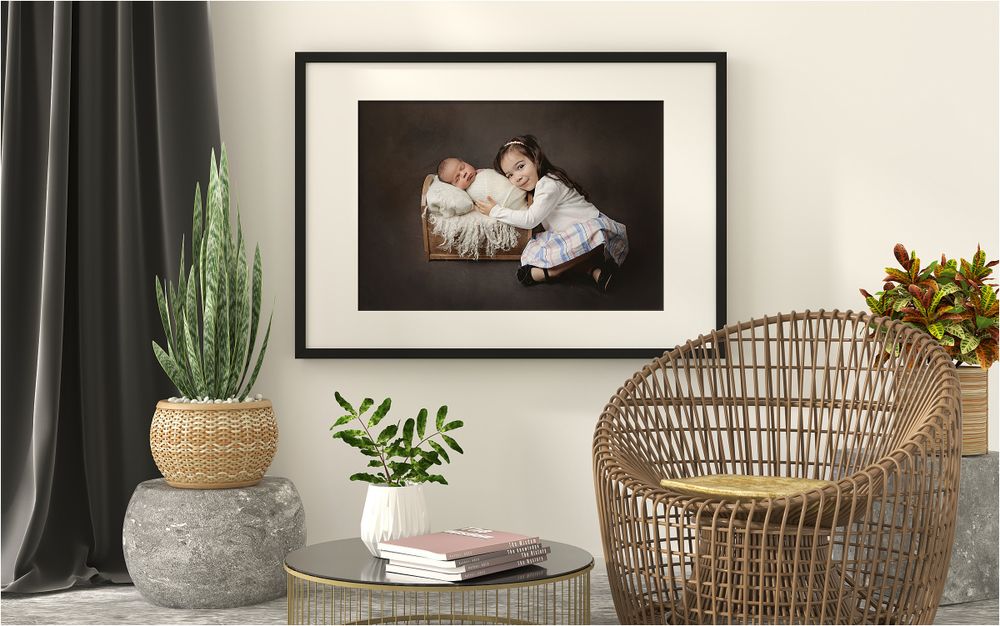 Framed newborn and sibling portrait displayed as wall art in room with plants and rattan chair