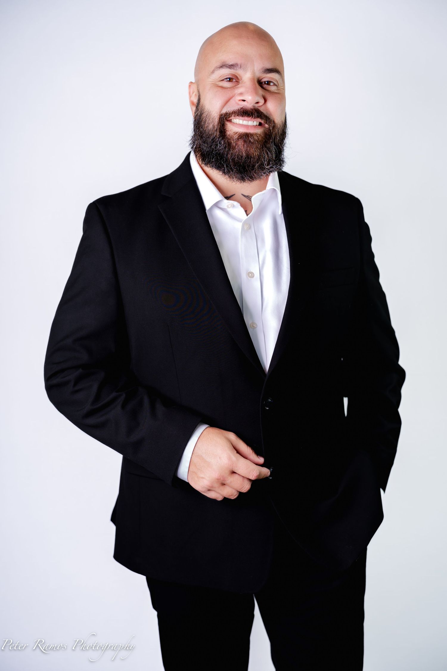 Professional portrait of a smiling person with a beard wearing a black suit jacket and white dress shirt against a light background.