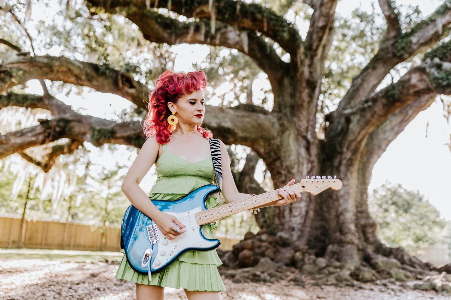 A musician with bright pink hair plays a blue electric guitar in front of ancient oak trees.