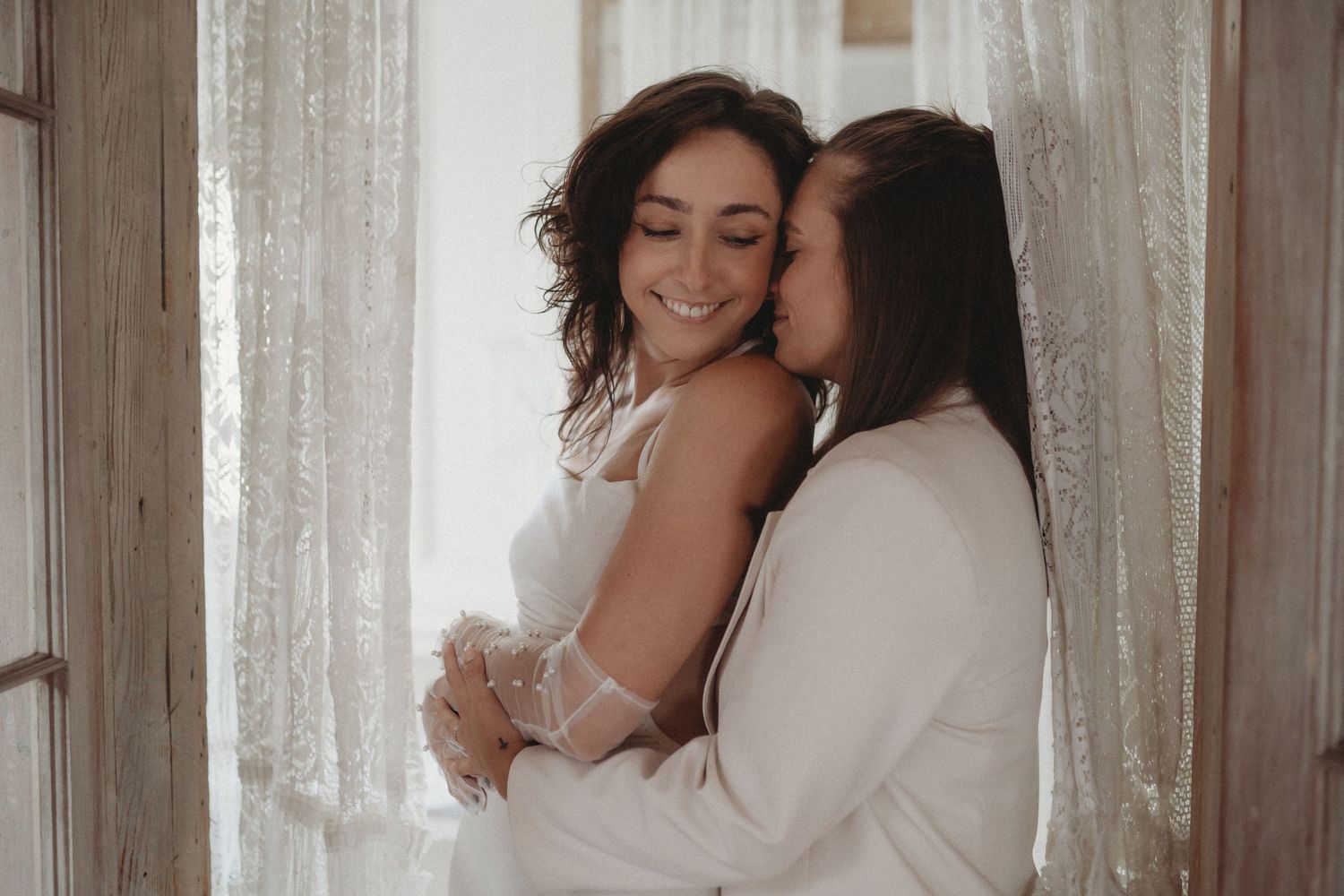 Intimate embrace captured in soft window light with white lace curtains.