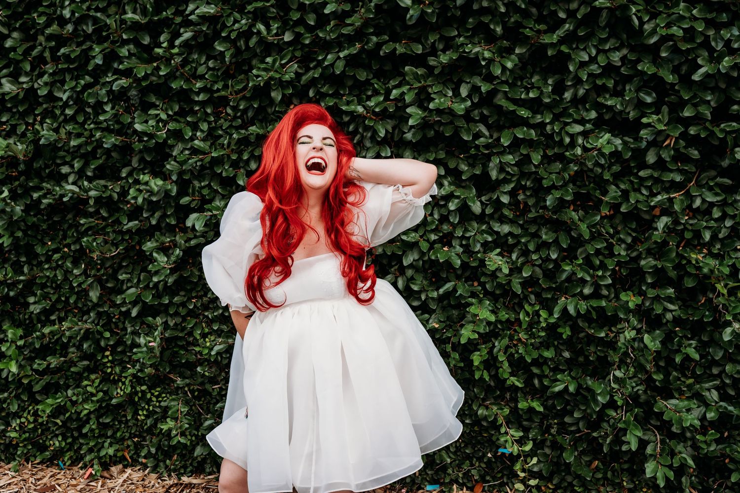 Woman in white dress with bright red hair laughing against hedge wall.