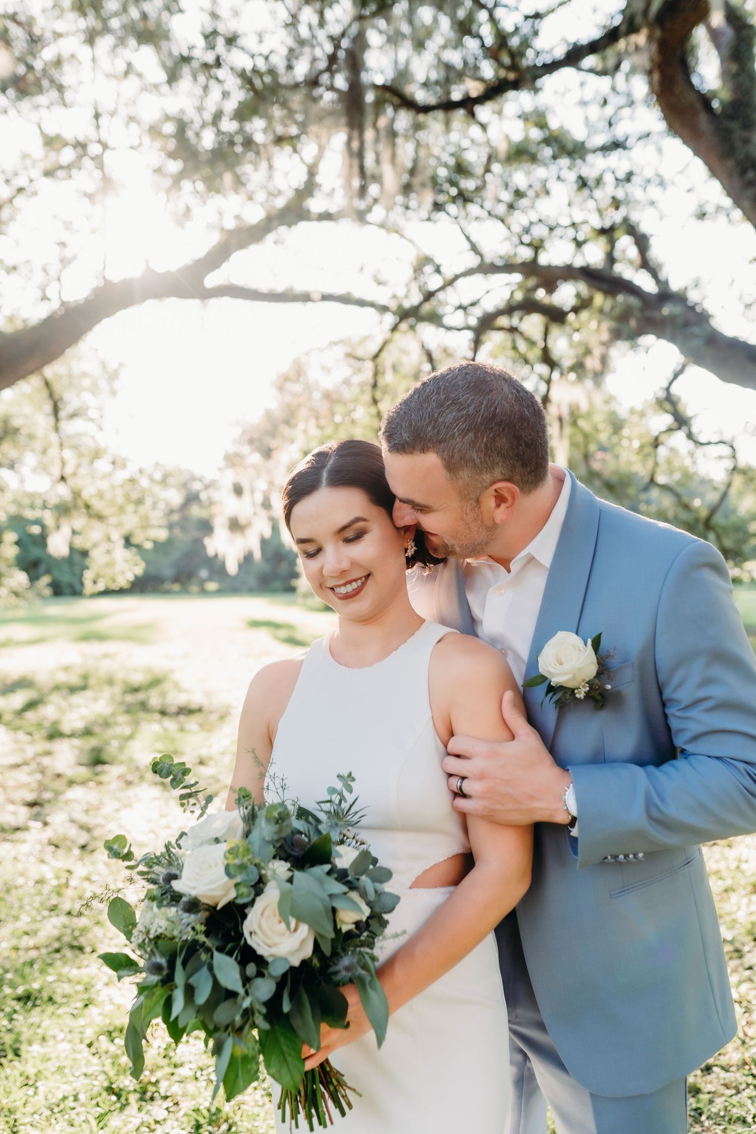 Tender moment between couple under sunlit trees with white bouquet.