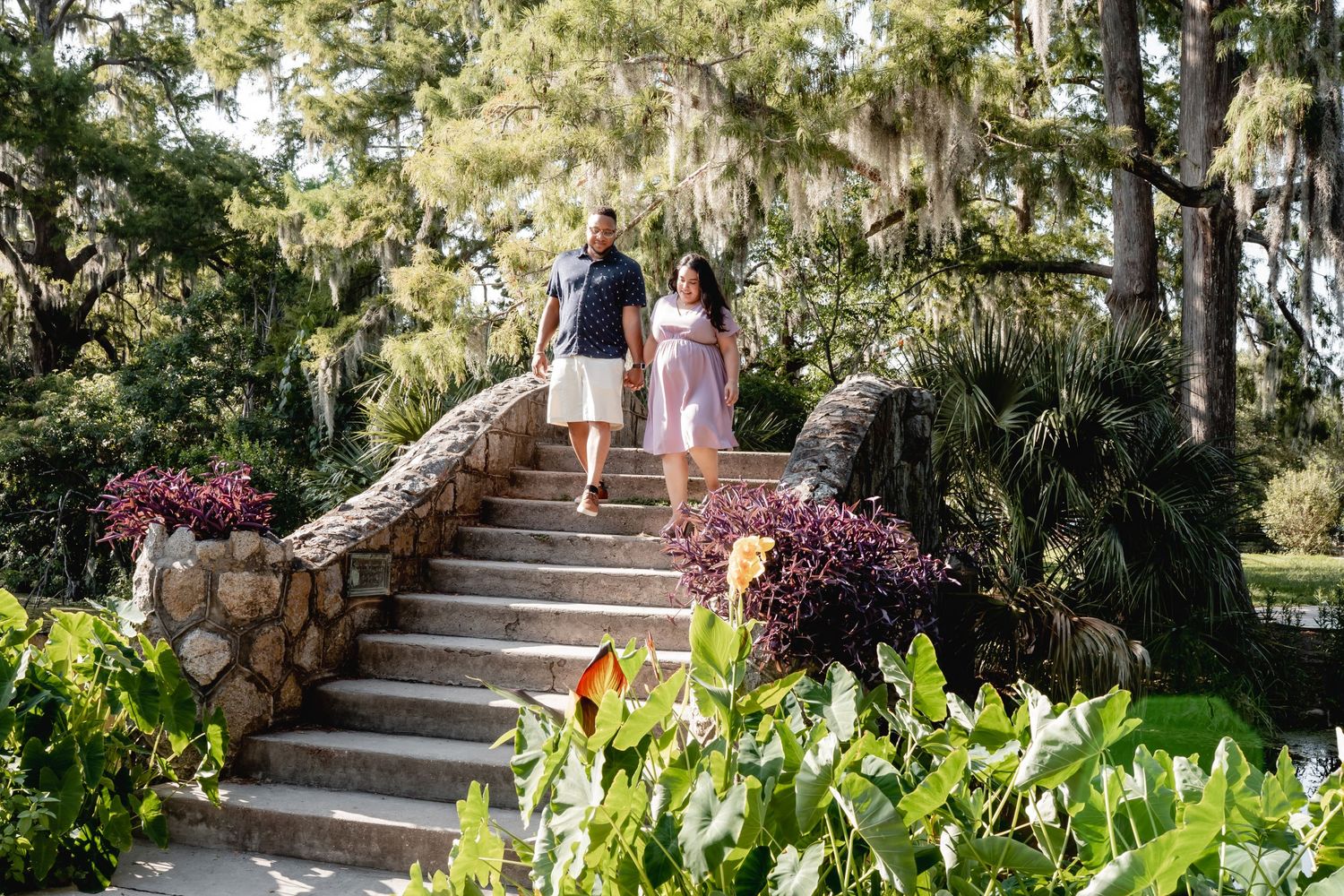 Couple ascending stone garden steps surrounded by blooming flowers and moss-draped trees.