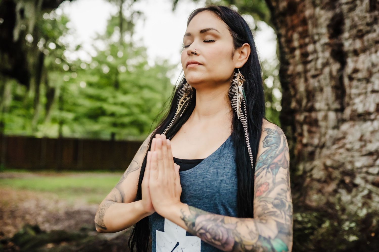 A meditative woman with tattoos and long dark hair practices mindful breathing outdoors near a tree.