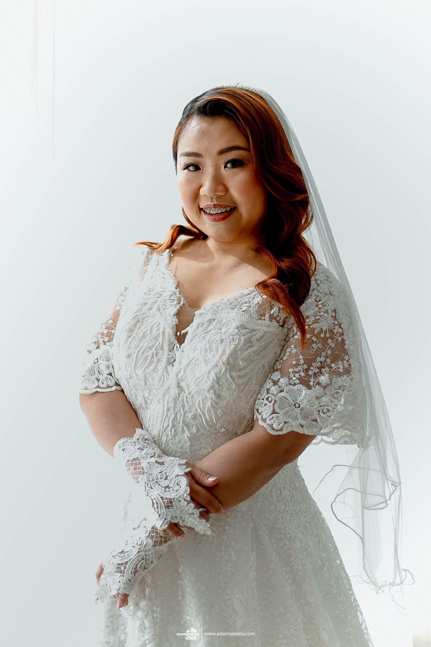 A bride poses in an elegant lace wedding gown with delicate sleeve details against a white background.