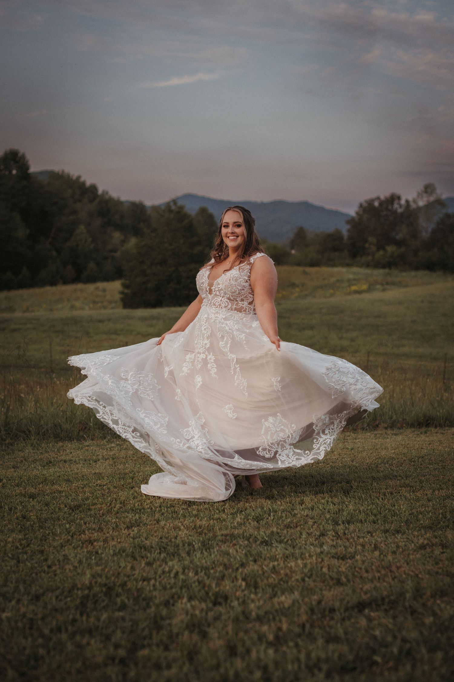 A flowing white wedding dress twirls in a grassy field at dusk with mountains in the background.