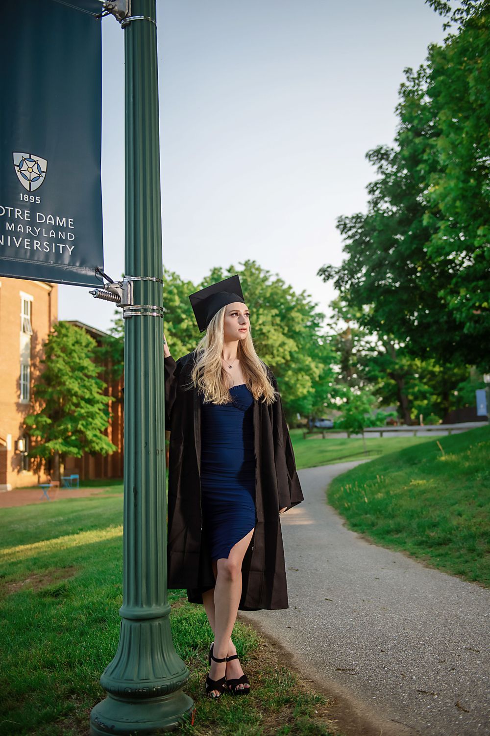 Graduate poses near green lamppost on campus pathway wearing cap and gown.