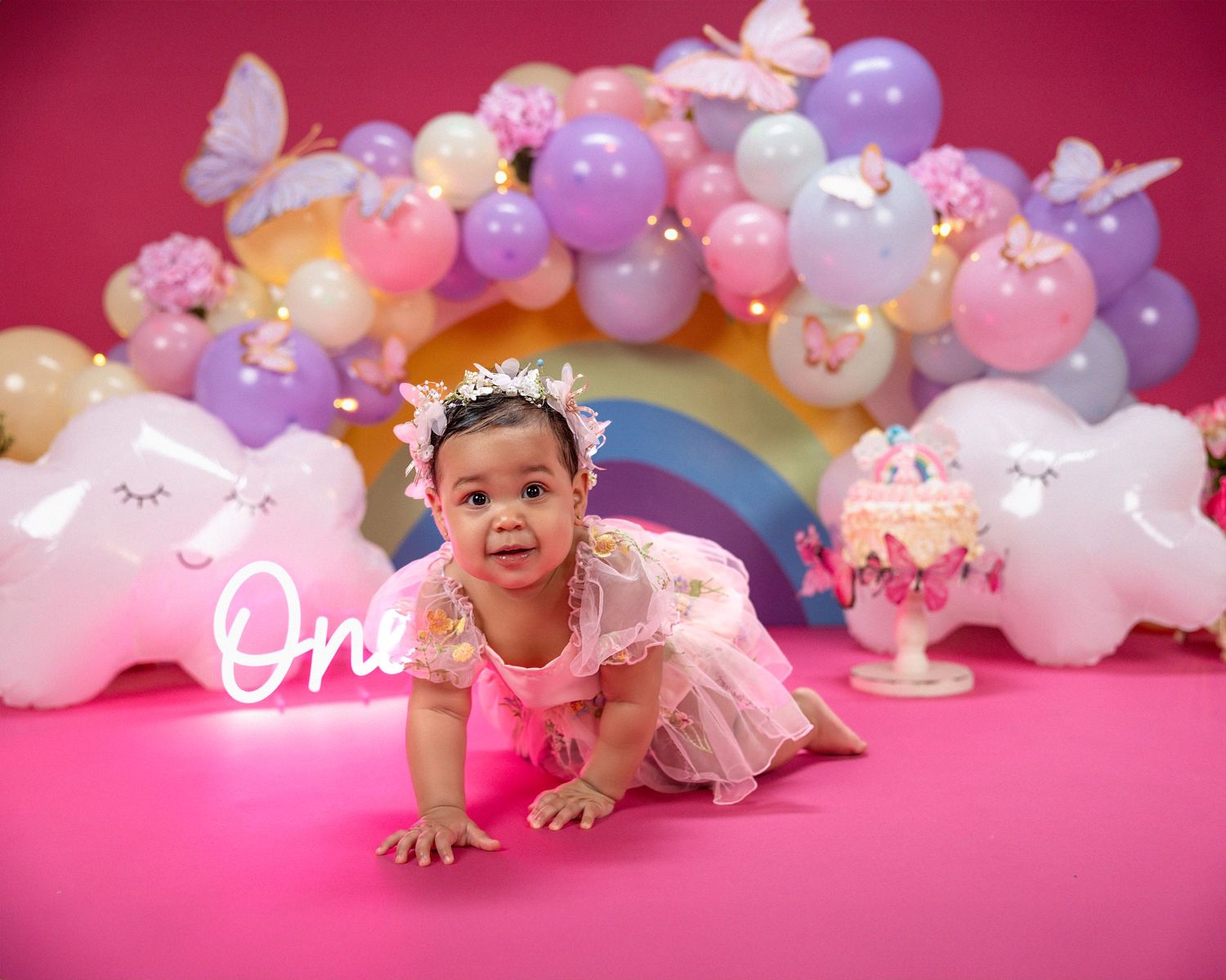 Baby in pink dress posing by colorful balloon arch with clouds and rainbow party decorations.