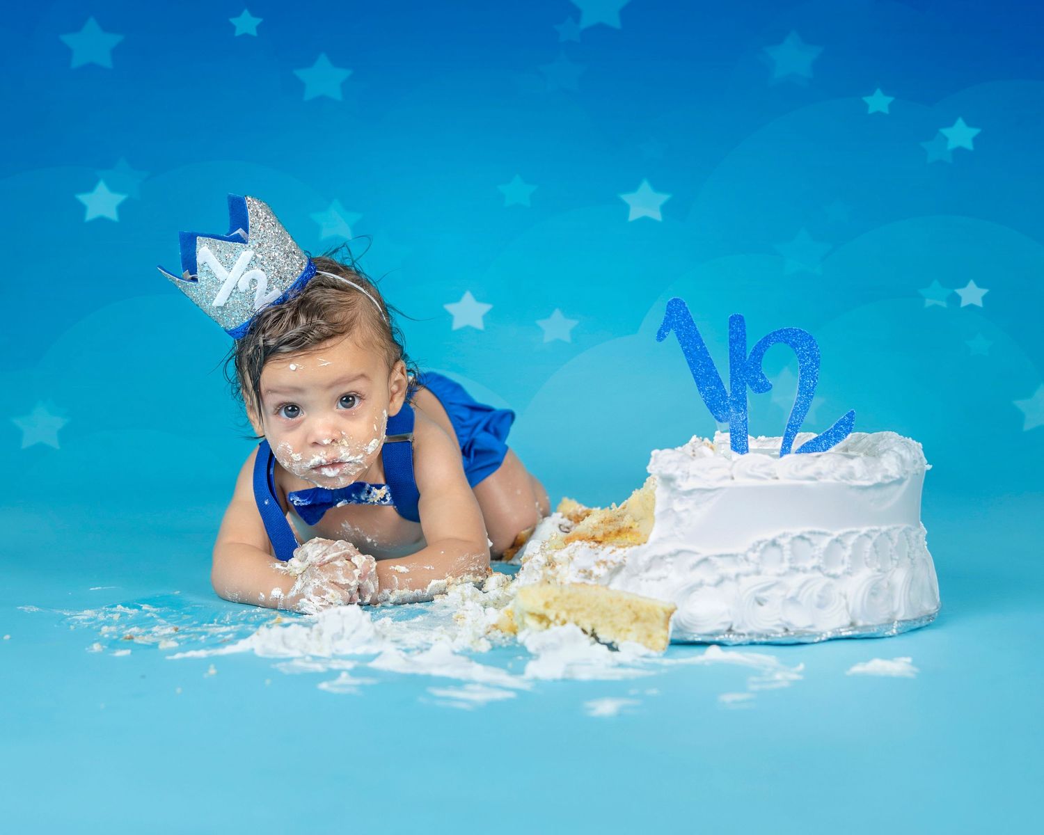 Baby in formal wear crawling next to smashed white birthday cake on blue background with stars.