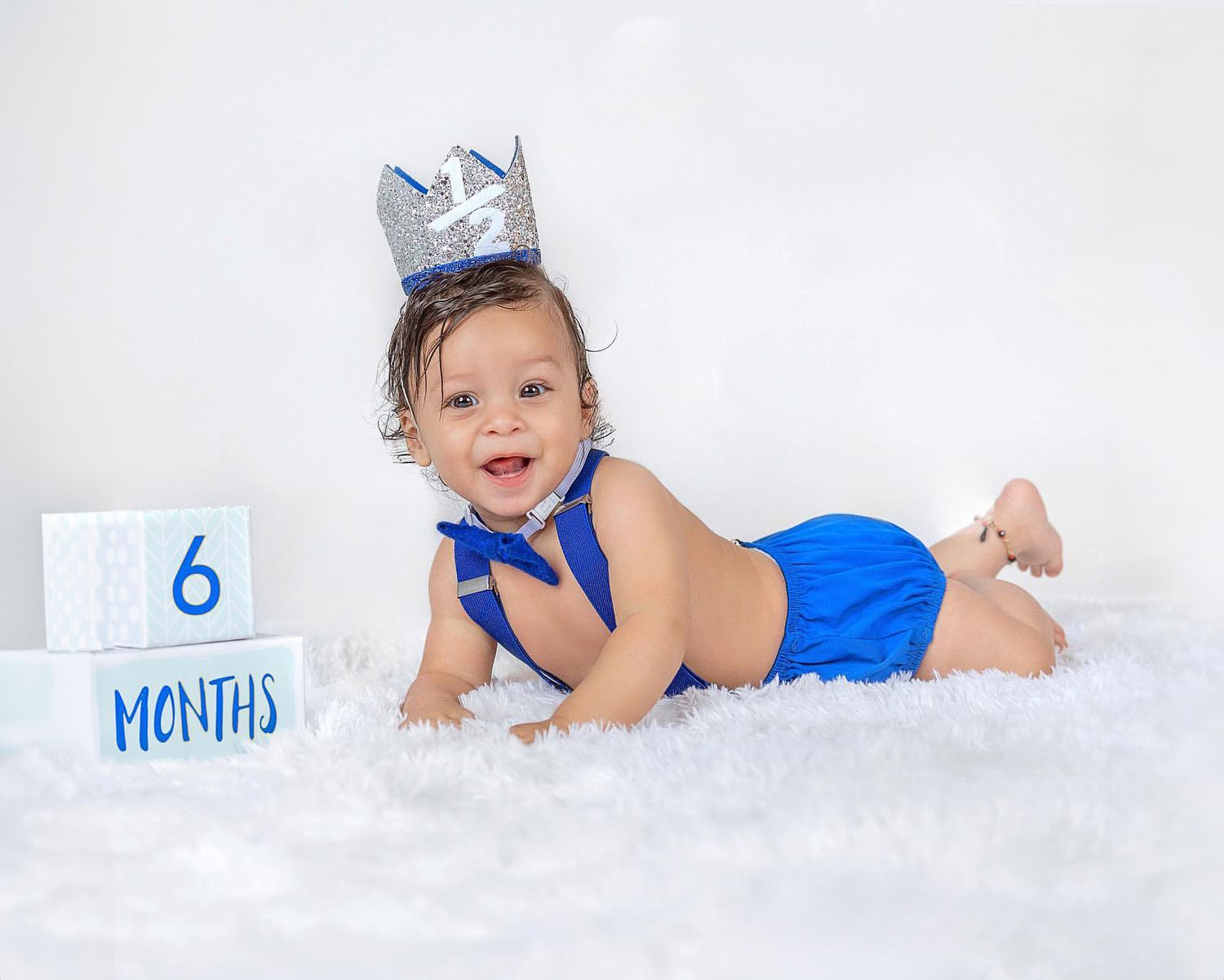 Baby milestone photo with silver crown and blue outfit on white fur backdrop.