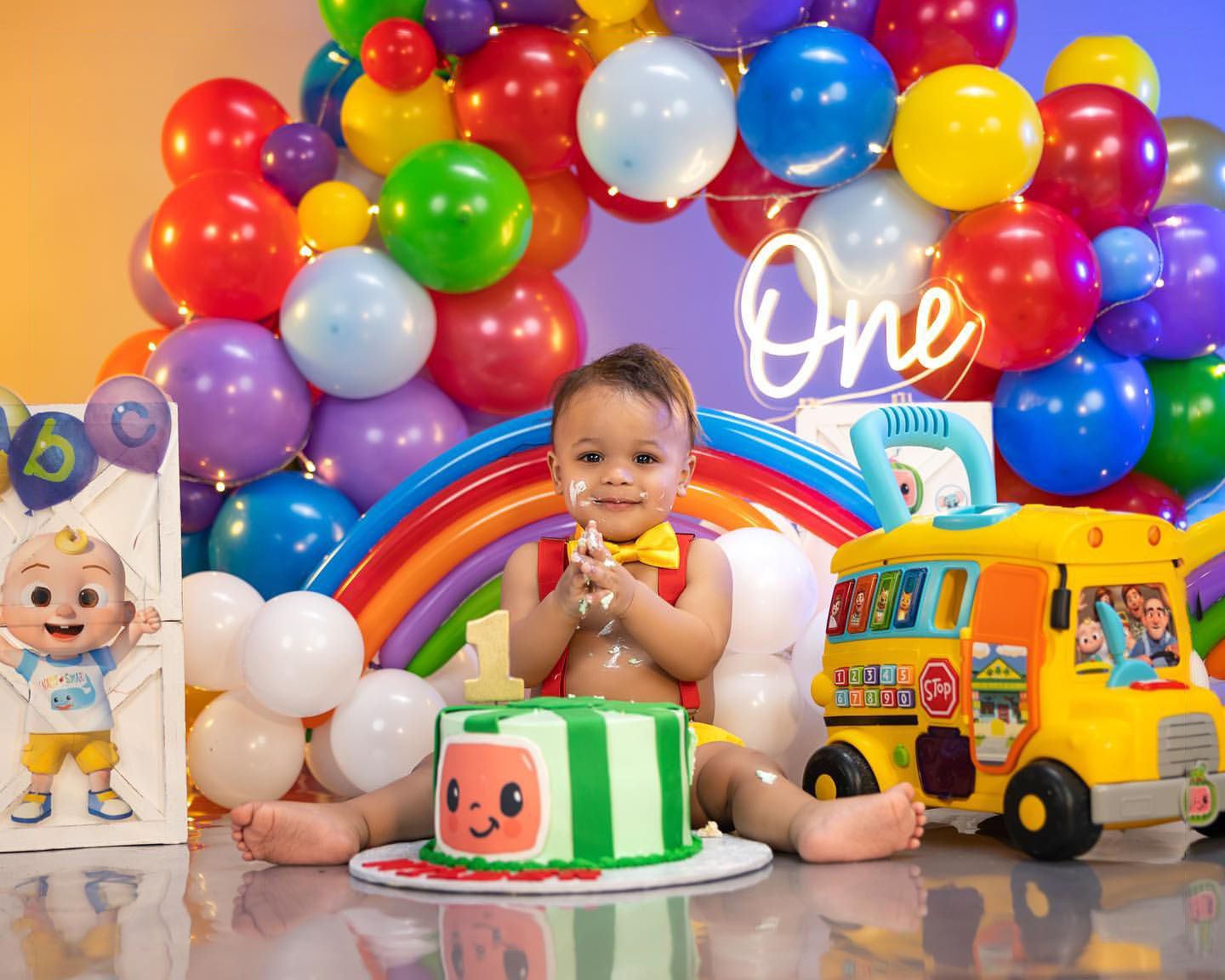 Rainbow balloon arch first birthday setup with colorful cake and toys.