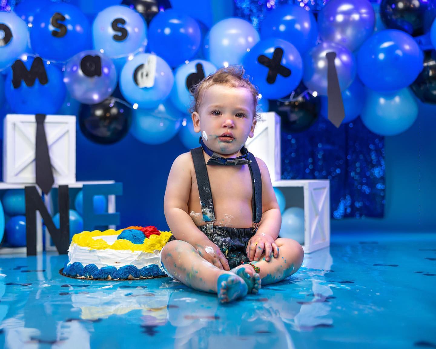 Blue and white balloon backdrop with seated baby in formal attire.