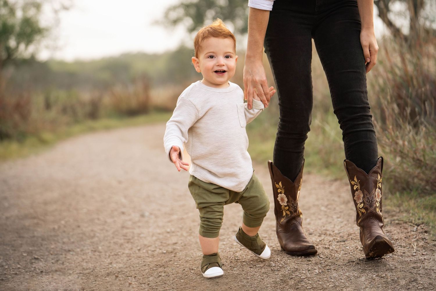 a mom in black pants and cowboy boots holds her toddler son's hand as they walk along a path at Phil Hardberger Park