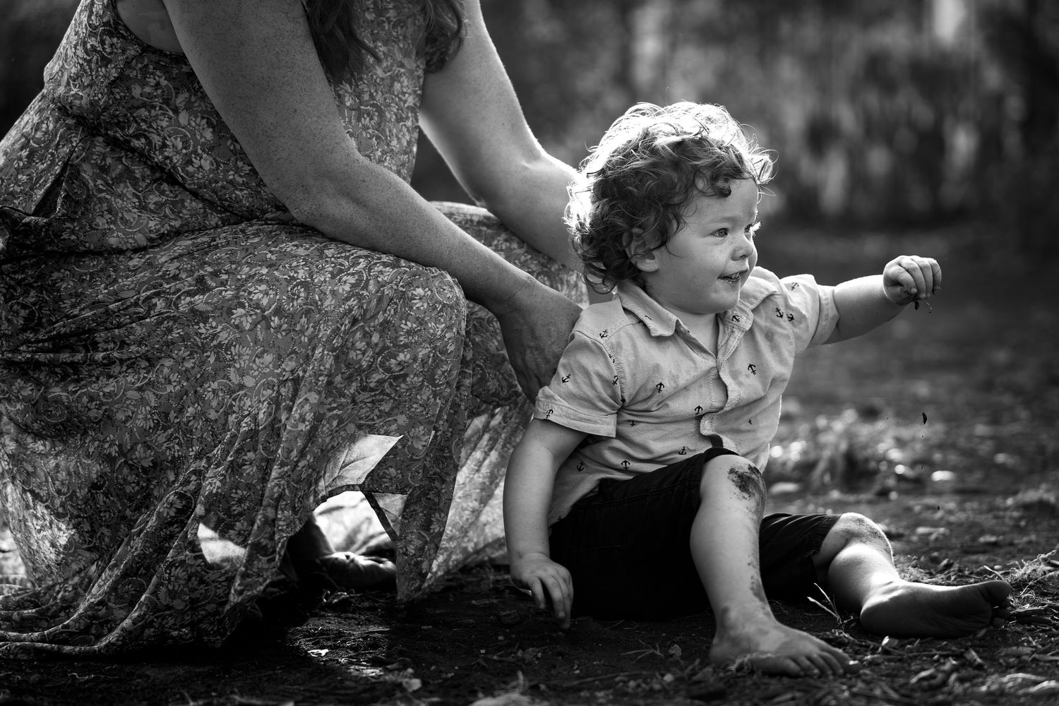 a black and white portrait of a toddler boy playing in the dirt with mom kneeling behind him