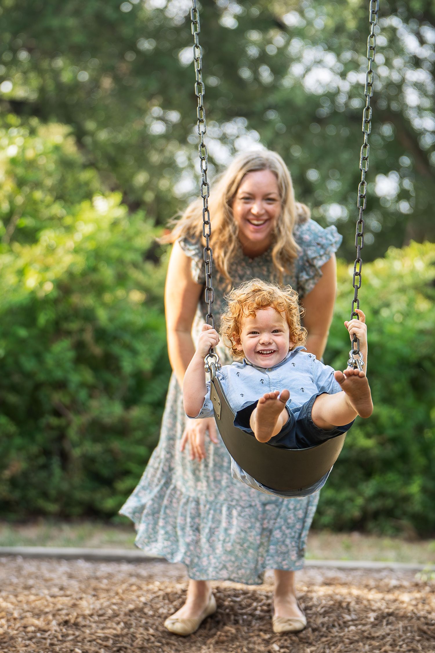 a mother wearing a long dress pushes her toddler son with red curly hair on a swing at Landa Library playground