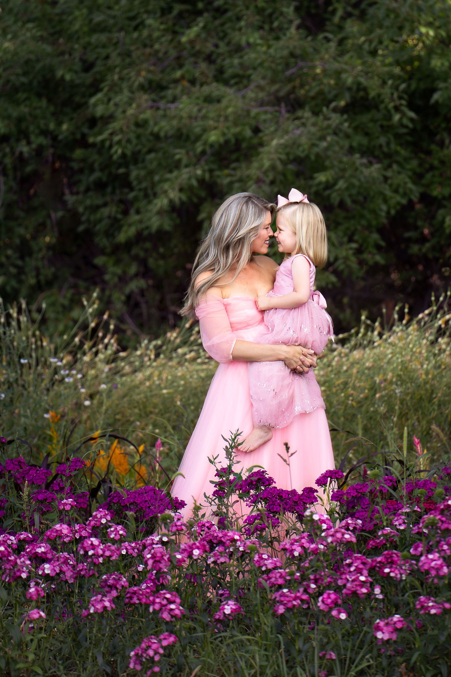 a mother wearing a long pink dress holds her toddler wearing a long pink dress in a field of purple and pink flowers