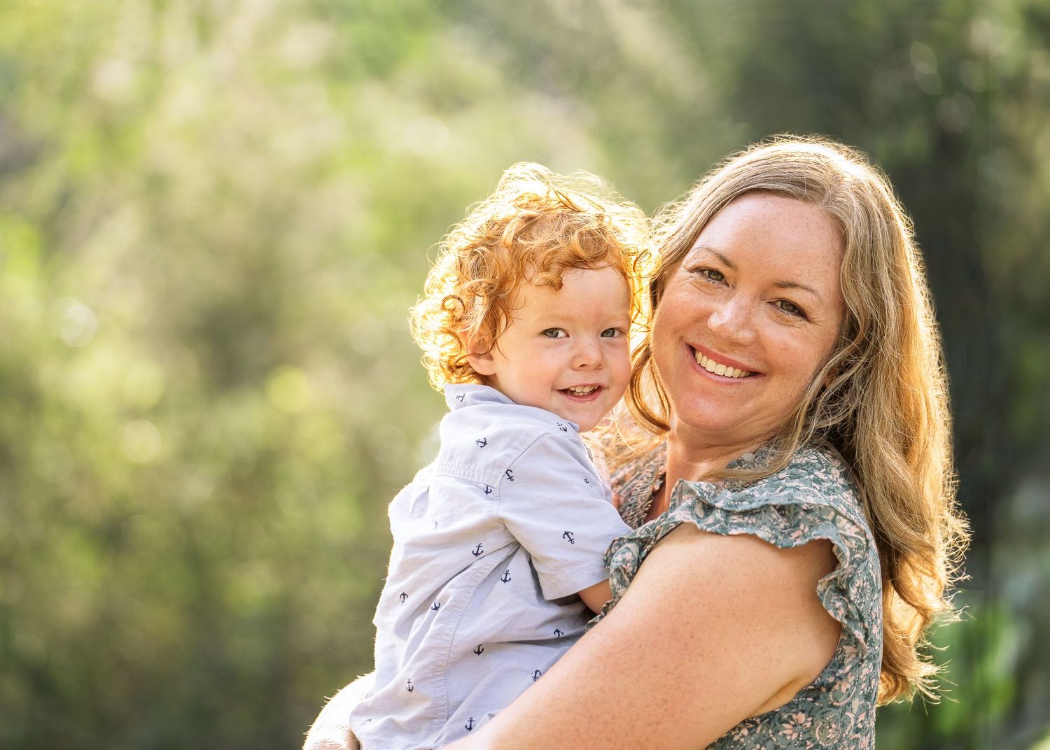 portrait of a mom holding her toddler son with red curly hair and sunshine glowing behind them while they smile