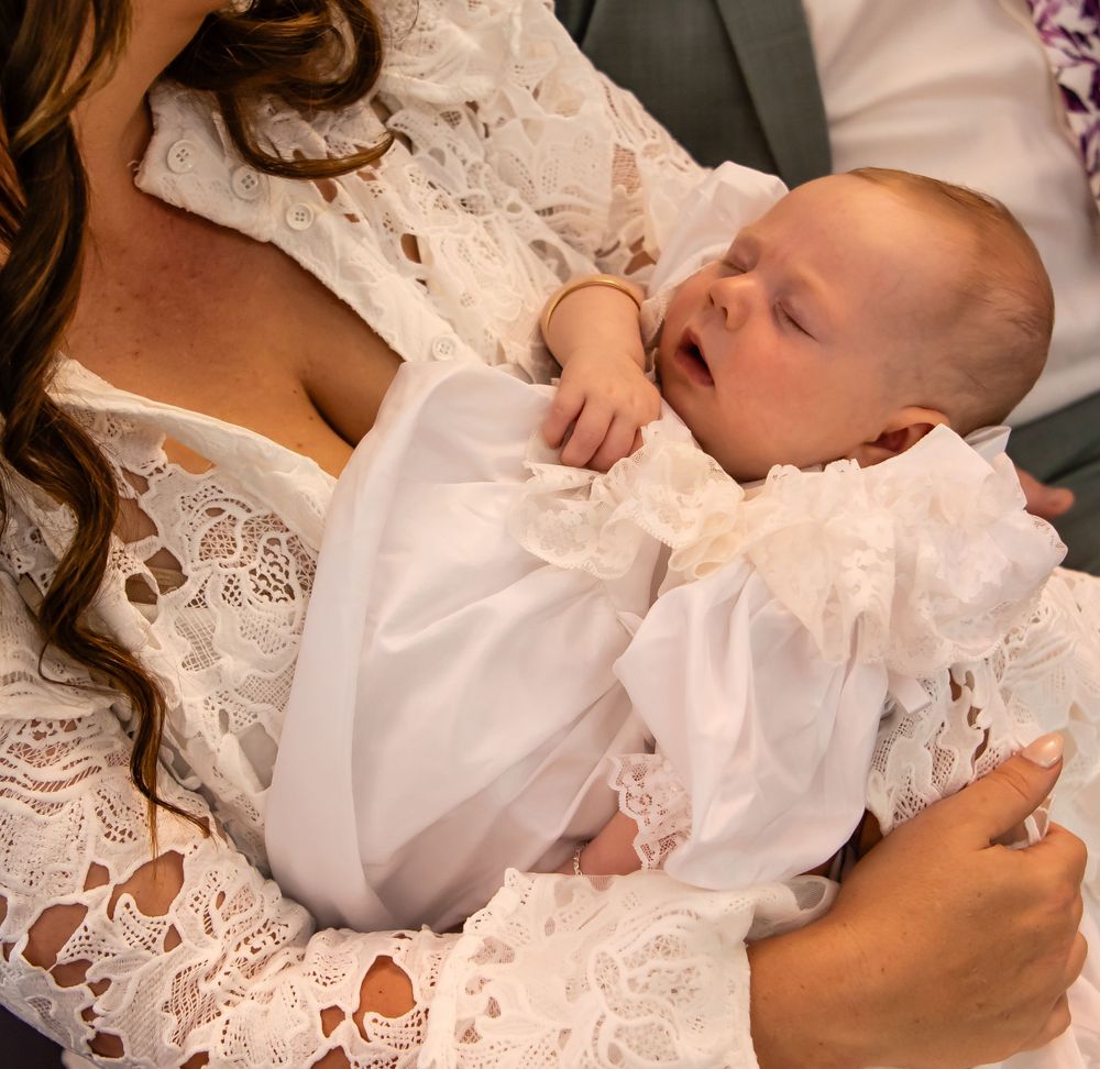 A sleeping newborn baby in white christening gown rests peacefully against white lace fabric.