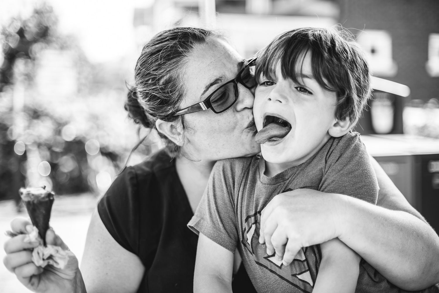 BW image of a mama giving her boy a big old kiss on the cheek as he sticks out his tongue.