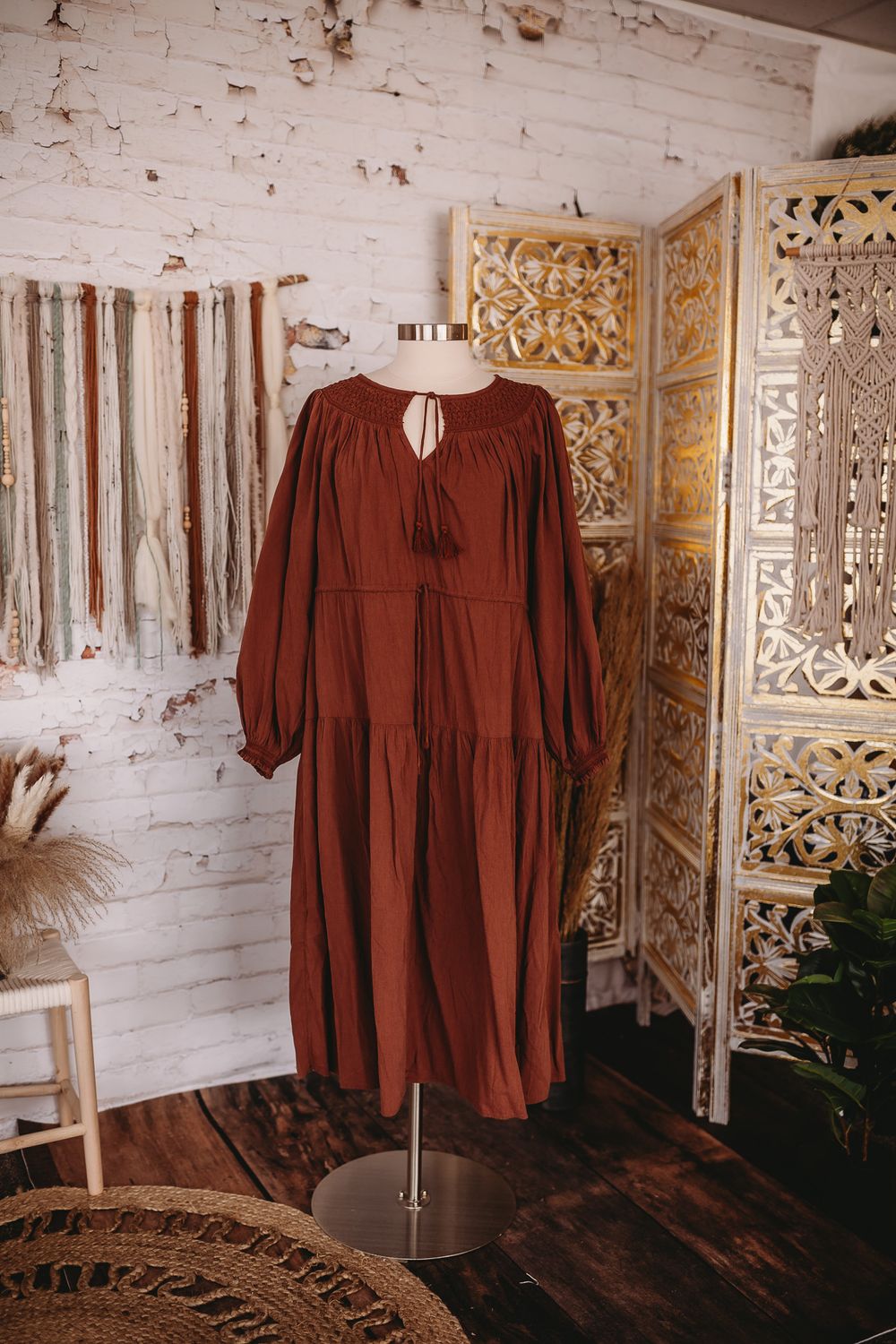 Rust brown tiered midi dress with tassel tie neckline and balloon sleeves on a mannequin in a boho studio.