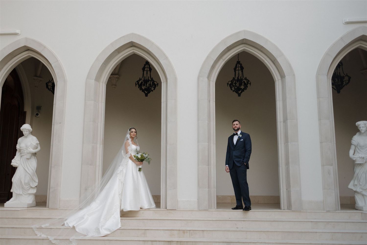 Wedding couple poses in gothic-style stone archways with classical statues.