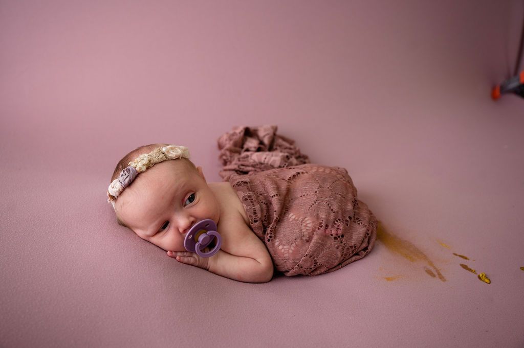 A sleeping baby with a pacifier wrapped in brown fabric against a pink background.
