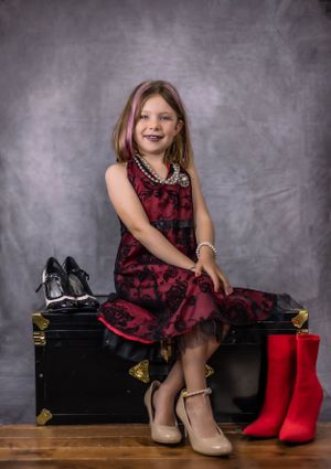 Someone in a burgundy dress poses playfully near boots on a black trunk.