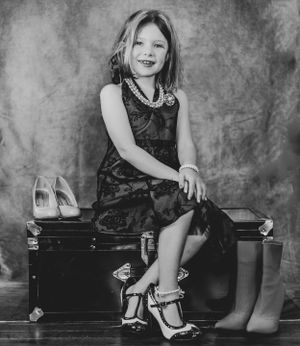 A portrait of someone in a dark dress sitting on a vintage trunk with dress shoes beside them.
