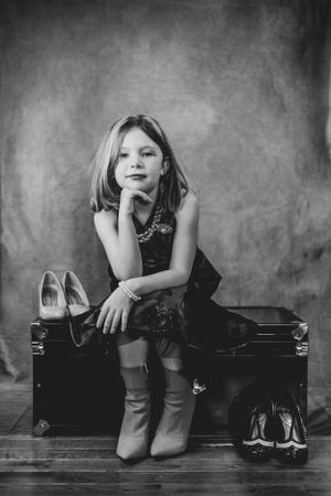 Black and white portrait showing someone seated on a trunk with shoes arranged nearby.