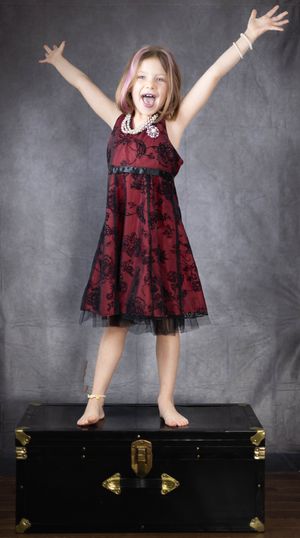 A joyful moment captured as someone in a burgundy dress jumps on a black trunk against a gray backdrop.