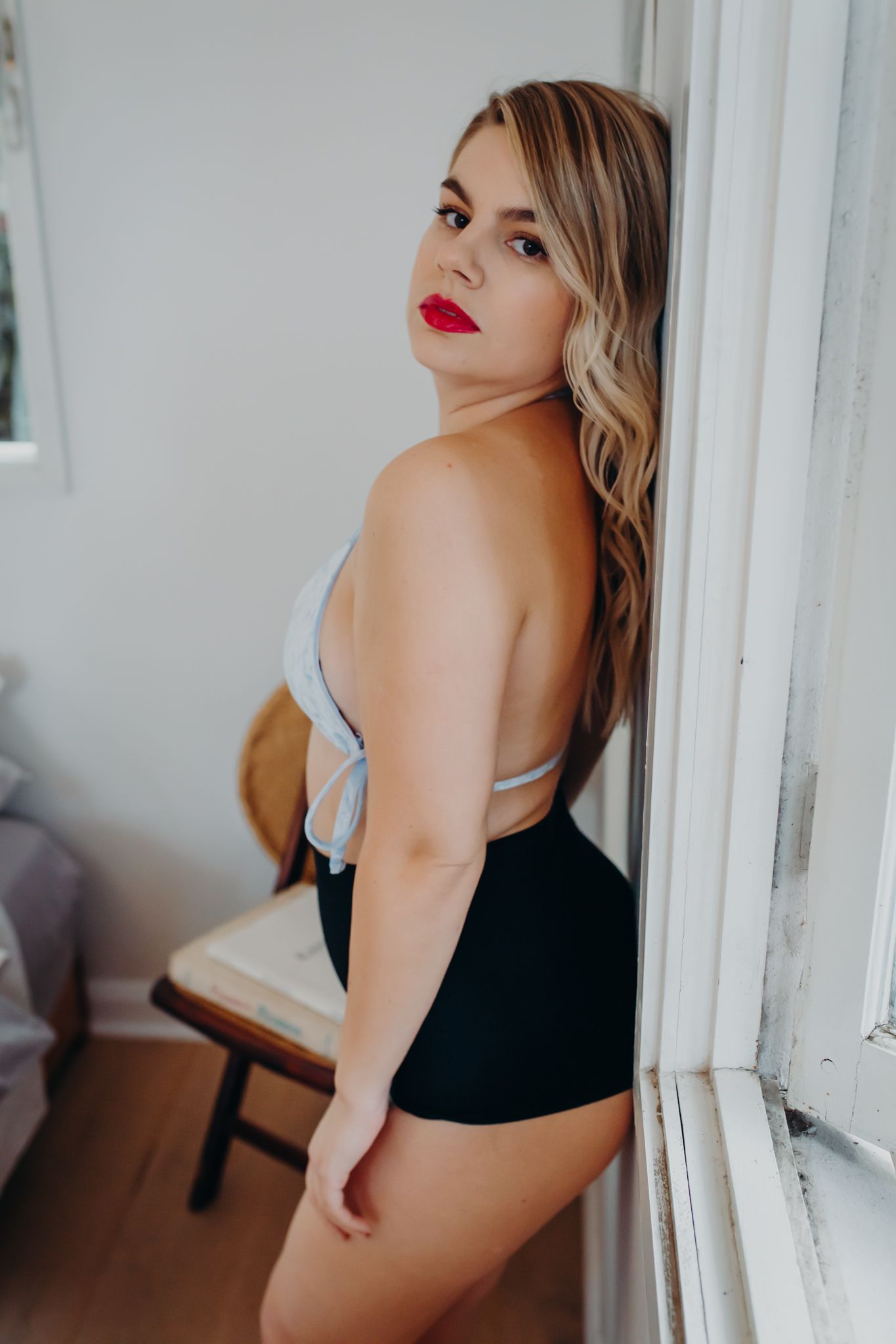 Model in black and gray lingerie poses seductively against a white wall in indoor boudoir photo.