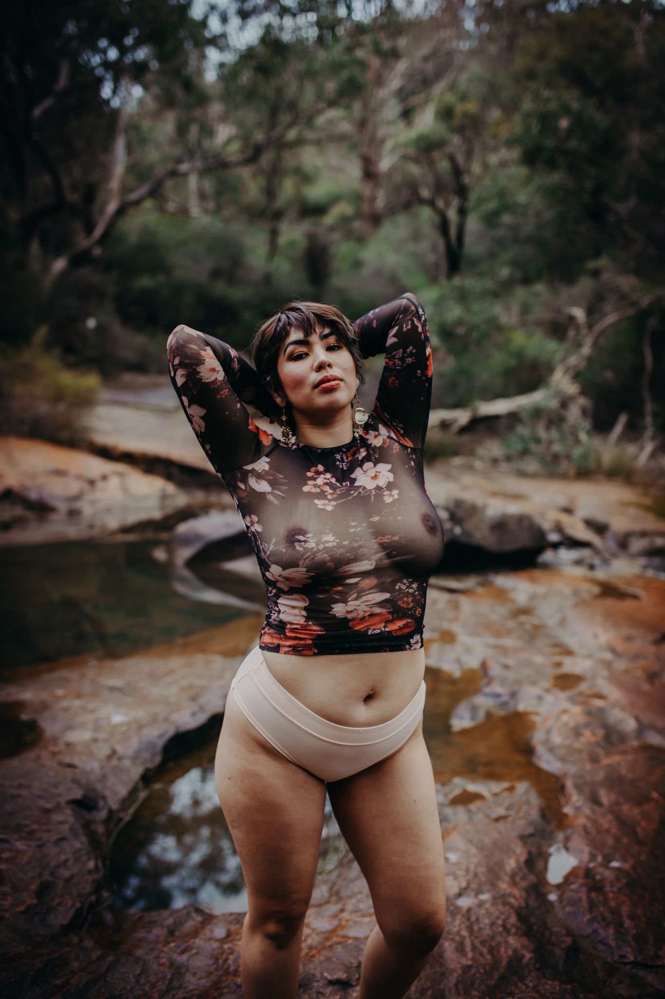 Model in sheer floral top poses at natural rock pools during a fashion shoot.