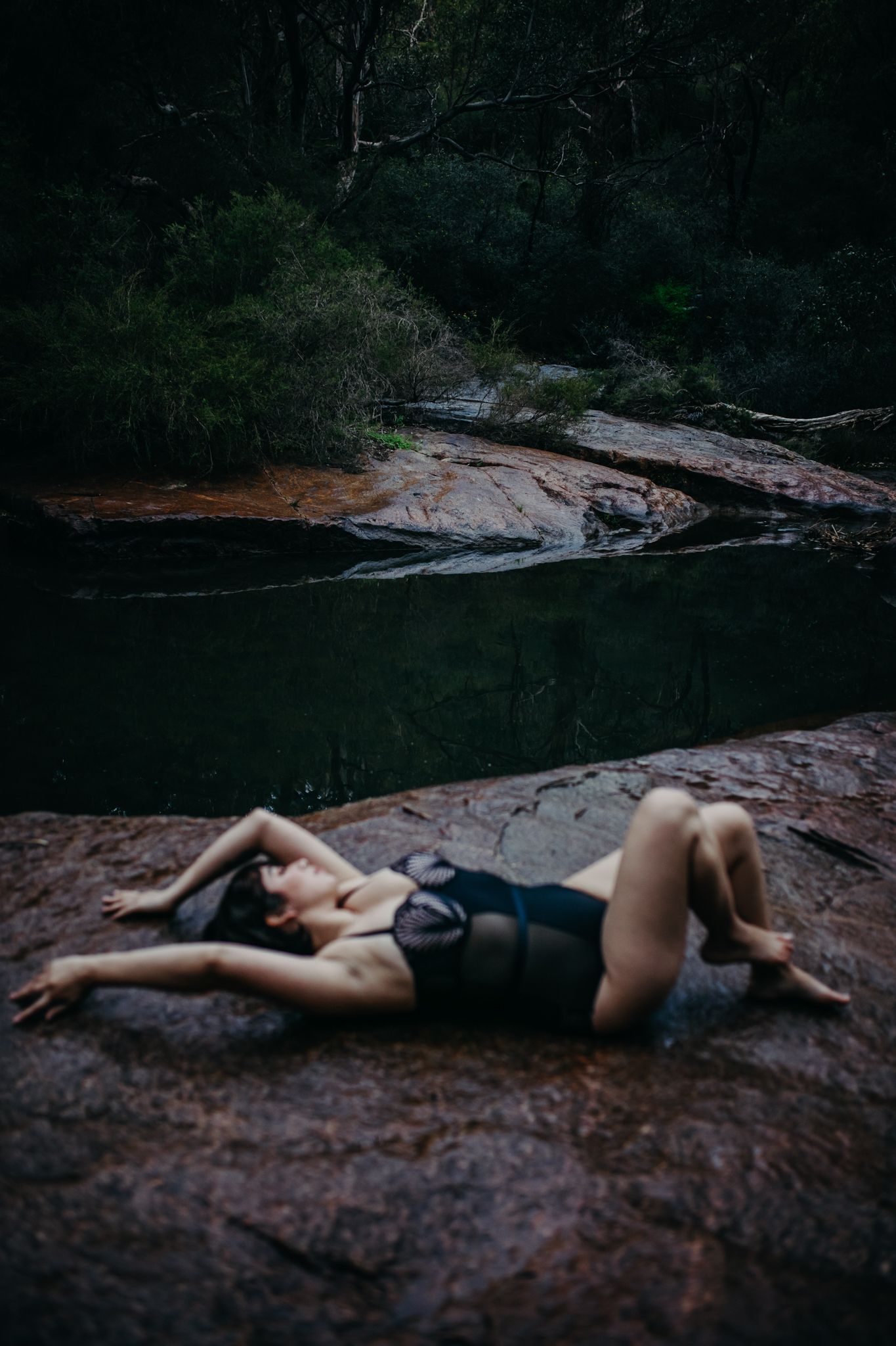 Artistic boudoir photo of a model reclining on rocks beside dark water.