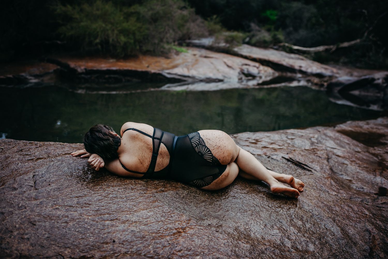 Model in black swimwear poses on red rocks near a serene water pool.