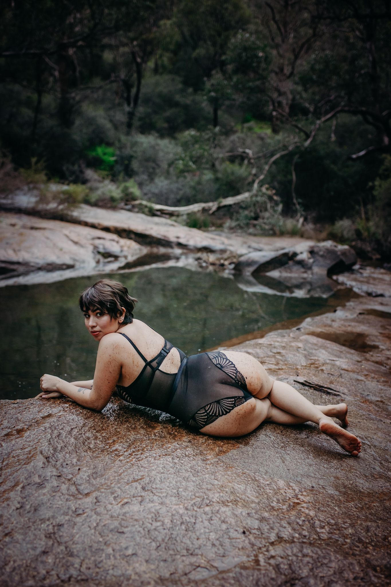 Model in black lingerie poses on red rock formations near a forest pool.