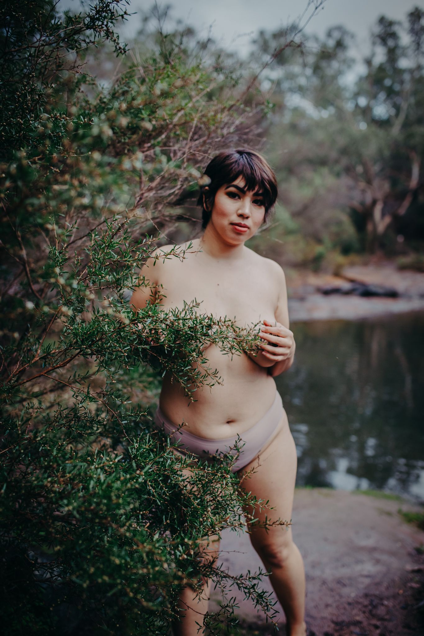 Natural boudoir portrait among leafy branches beside a tranquil pond.