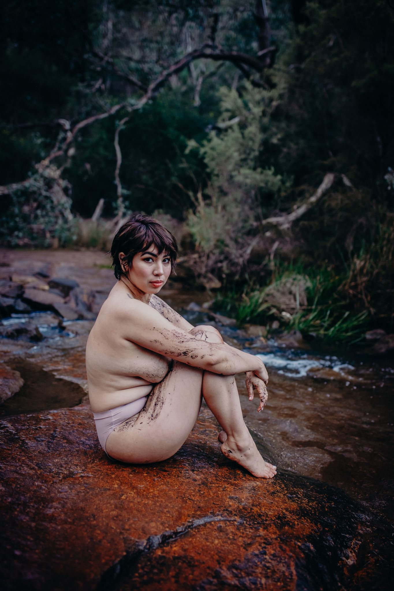 Minimalist outdoor portrait on red rocks beside a forest stream.
