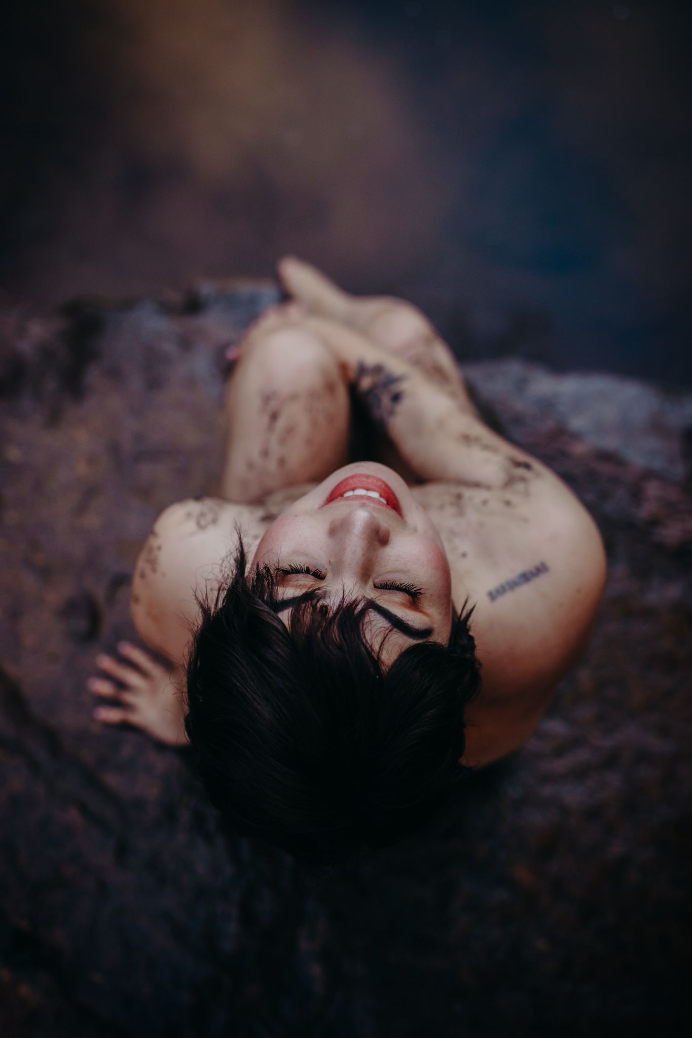 Artistic high angle view of someone lying down covered in mud or soil with moody dark lighting and bokeh effects.