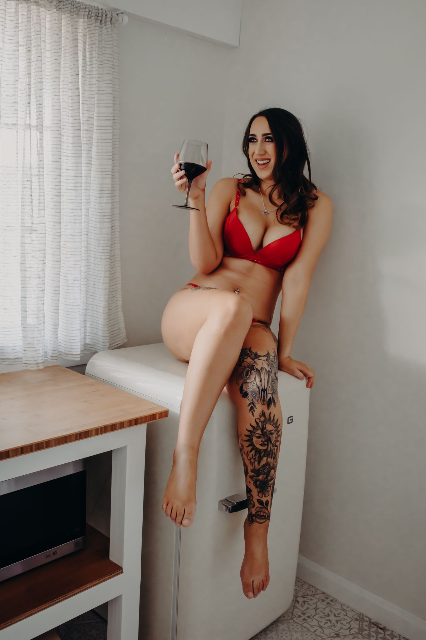 Boudoir photo of someone in red lingerie sitting on white furniture with a glass of wine and floral leg tattoo.