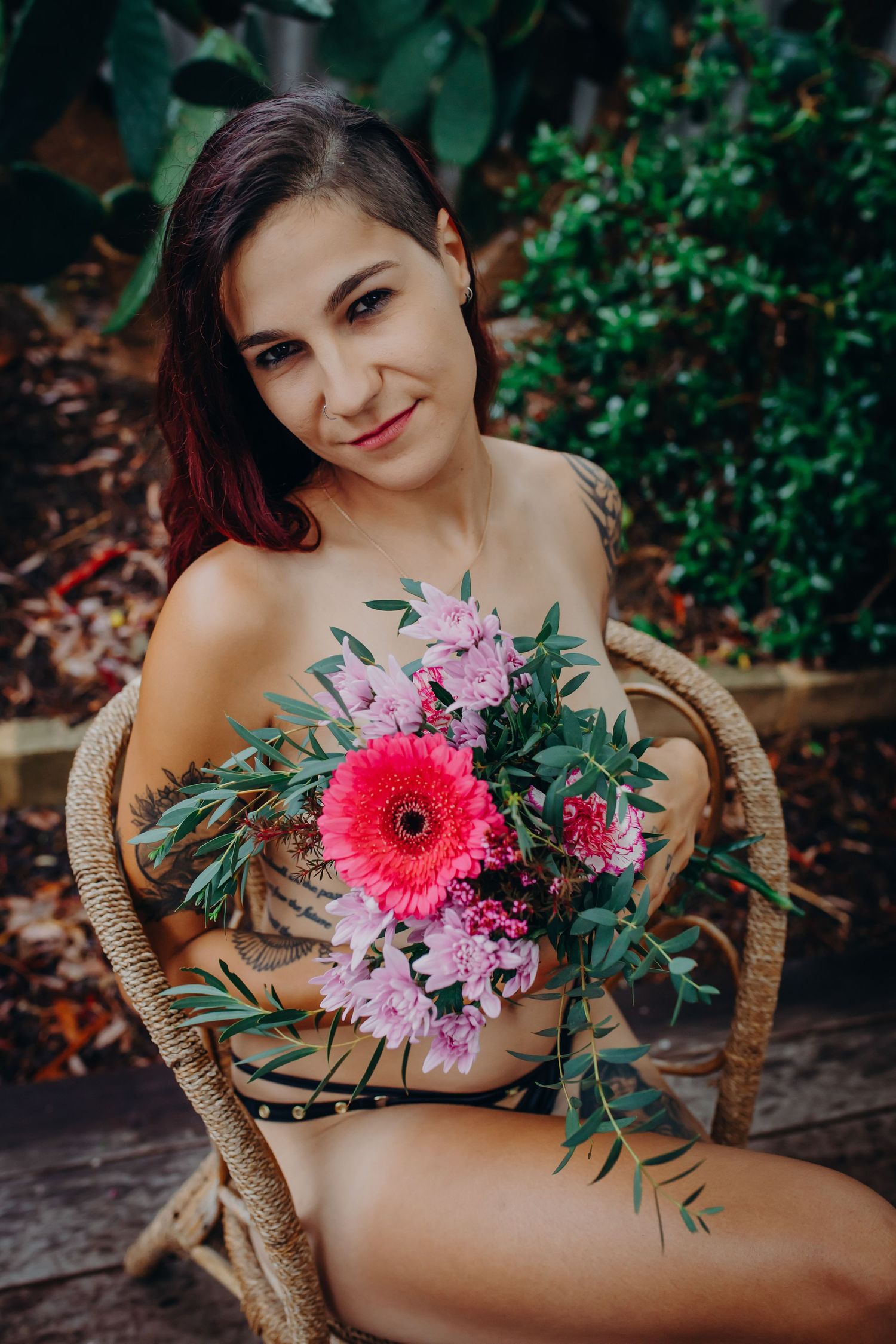 Model holding vibrant floral bouquet with pink gerbera daisy while sitting in wicker chair outdoors.
