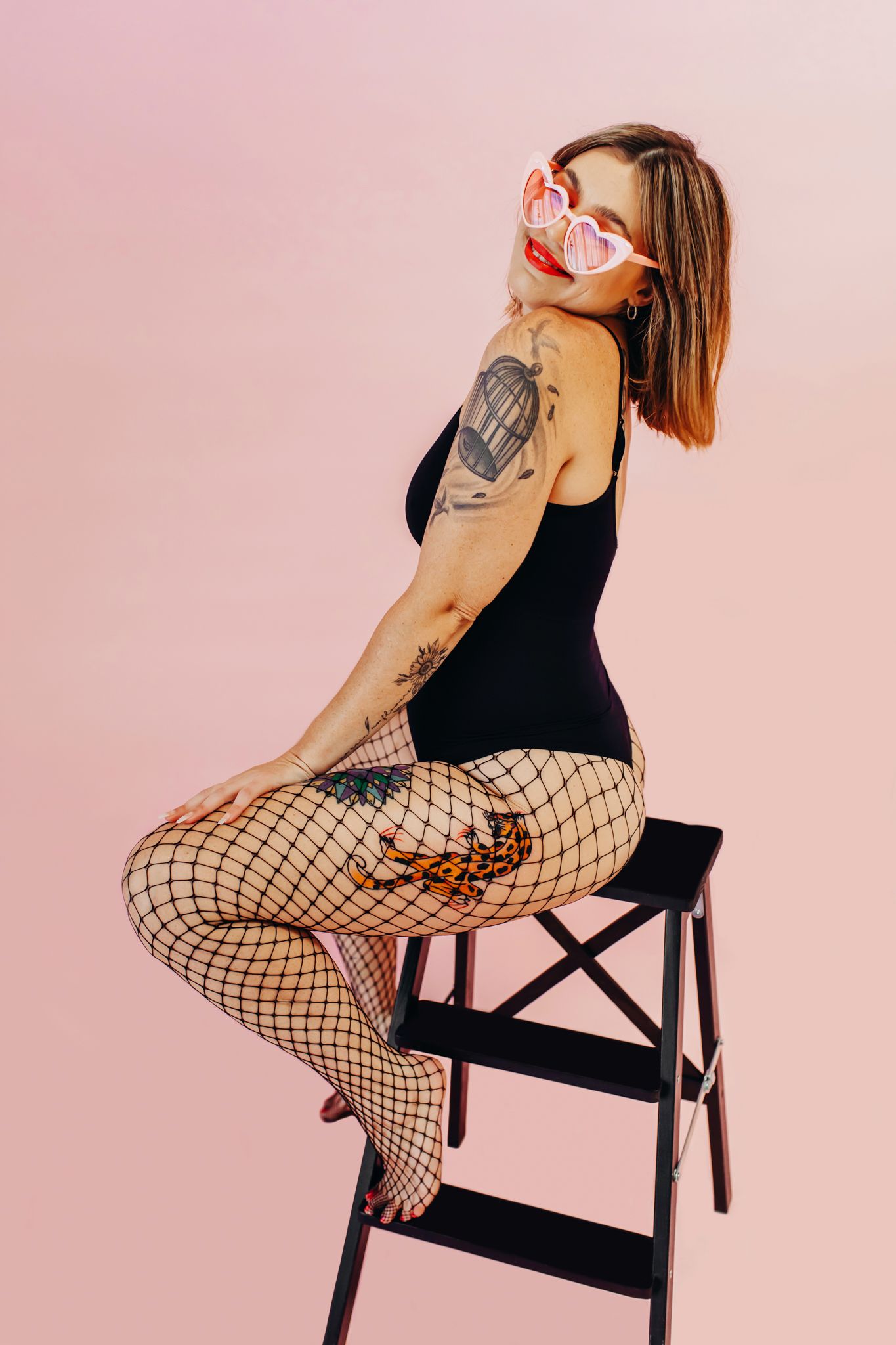 Person in black bodysuit and fishnet stockings posing on ladder against pink background.
