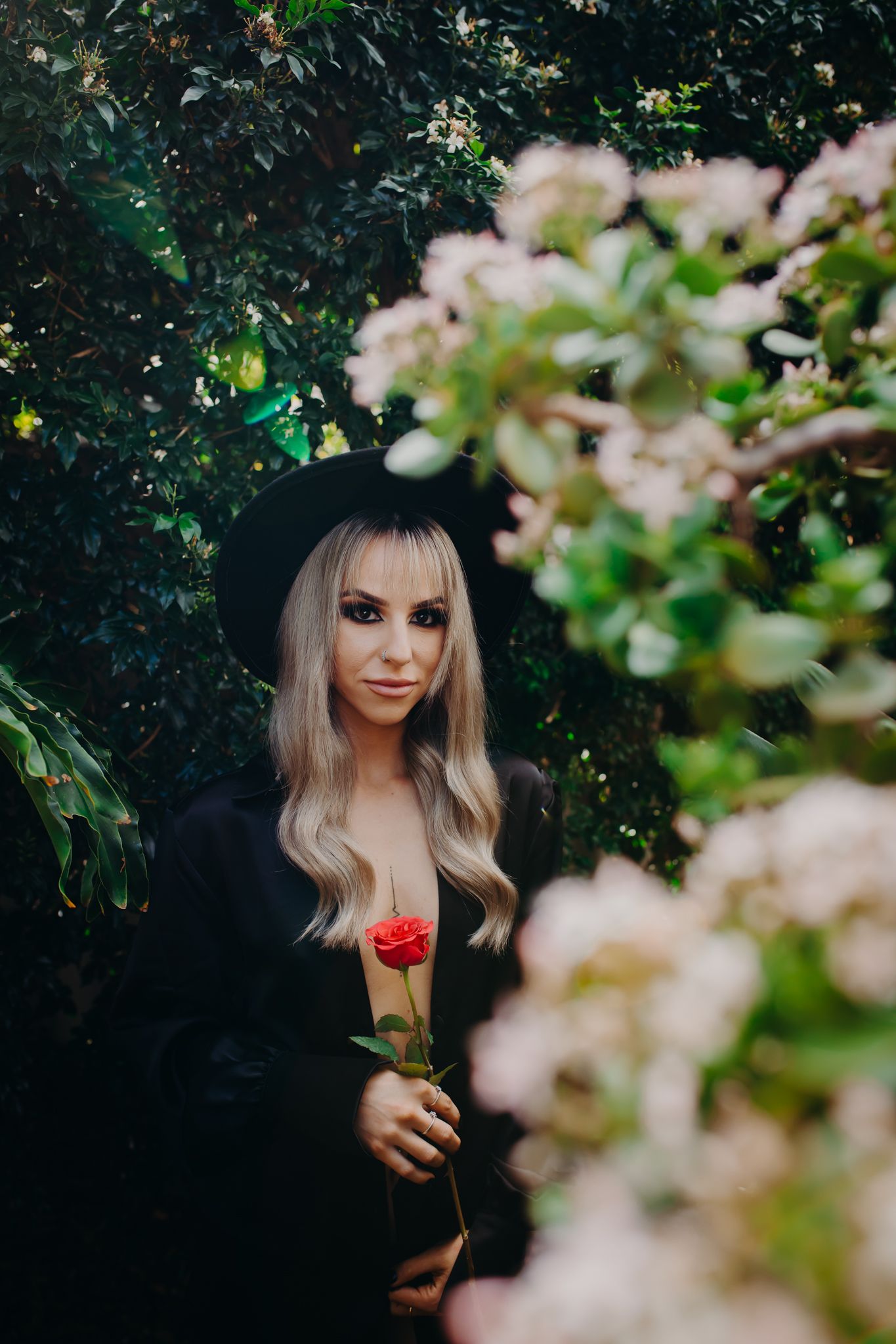 Ethereal portrait in black outfit holding red rose with soft cherry blossoms in foreground.