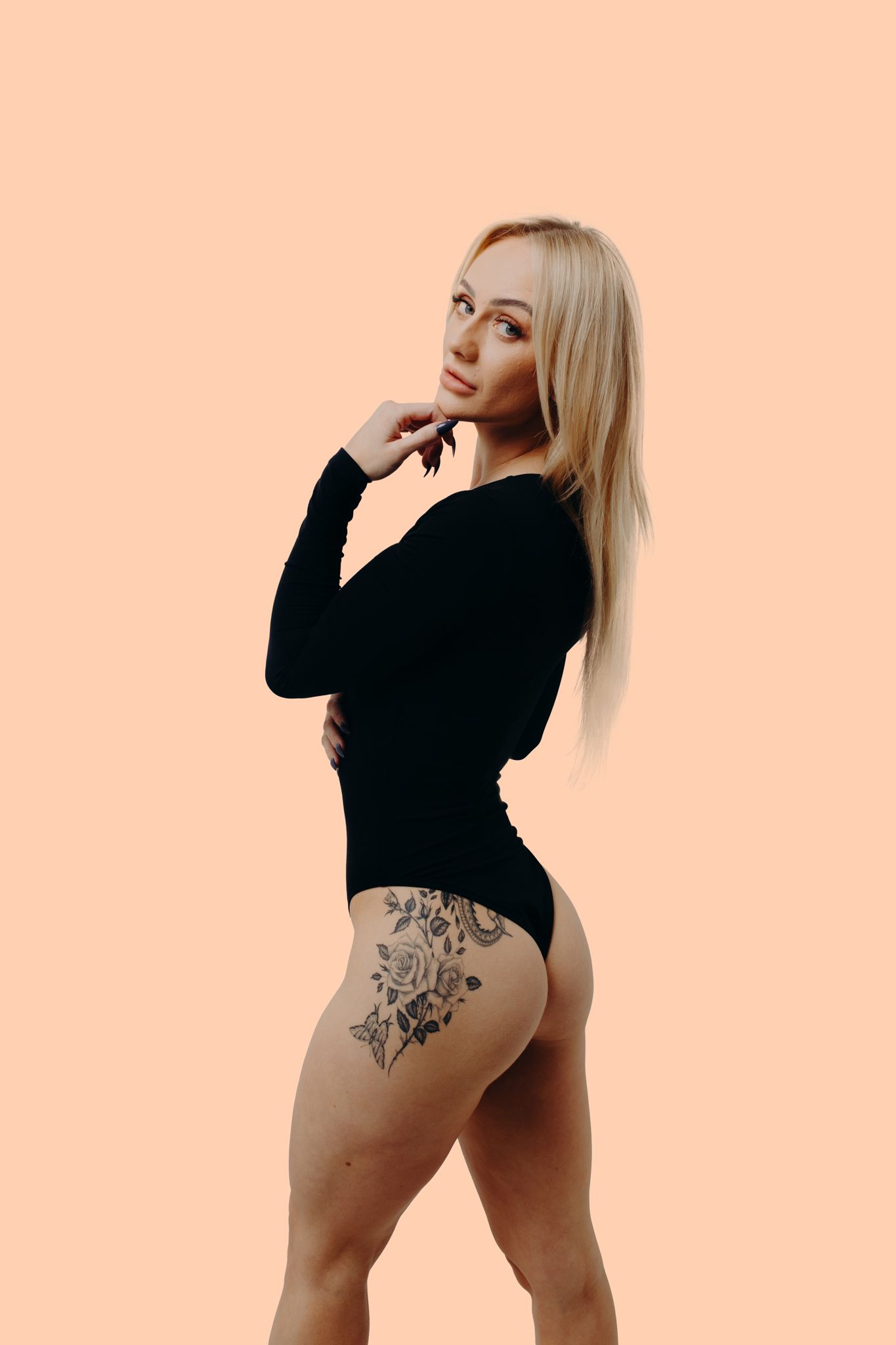 Athletic figure in black bodysuit poses against peach background showing floral hip tattoo.