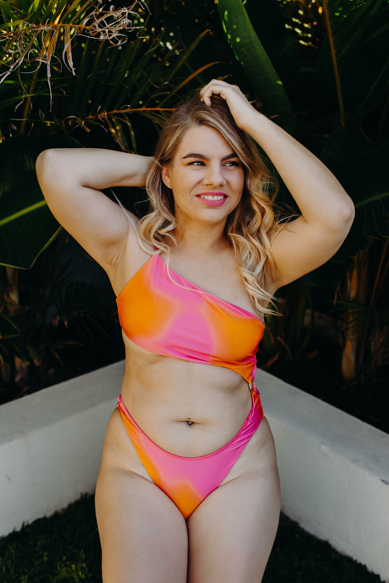 Confident pose in vibrant orange and pink gradient swimsuit against tropical backdrop.