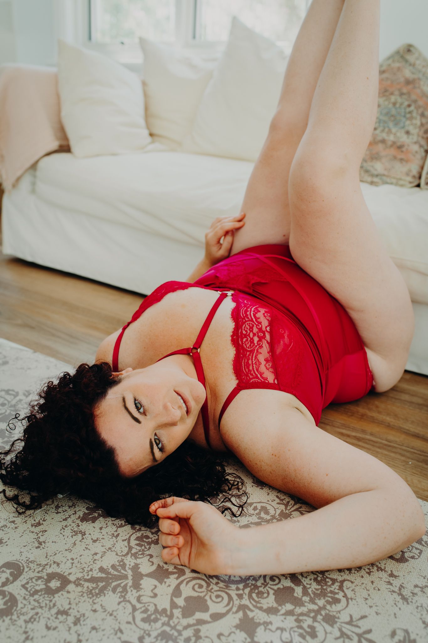 Model in red lace lingerie reclines on hardwood floor near white sofa.