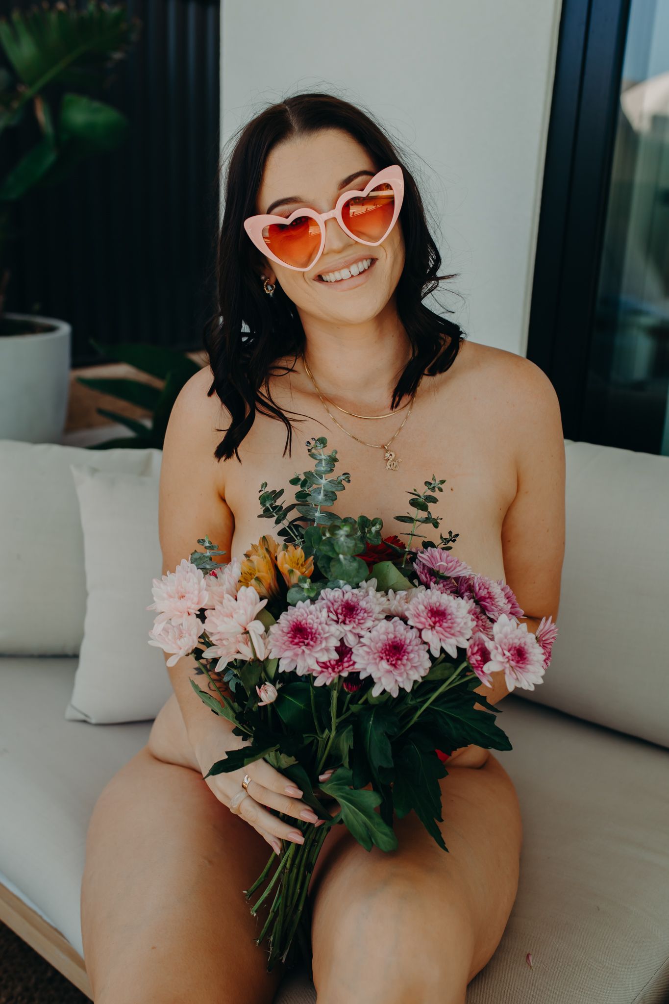 Person wearing heart-shaped sunglasses holding a colorful bouquet of flowers while sitting on a white couch.