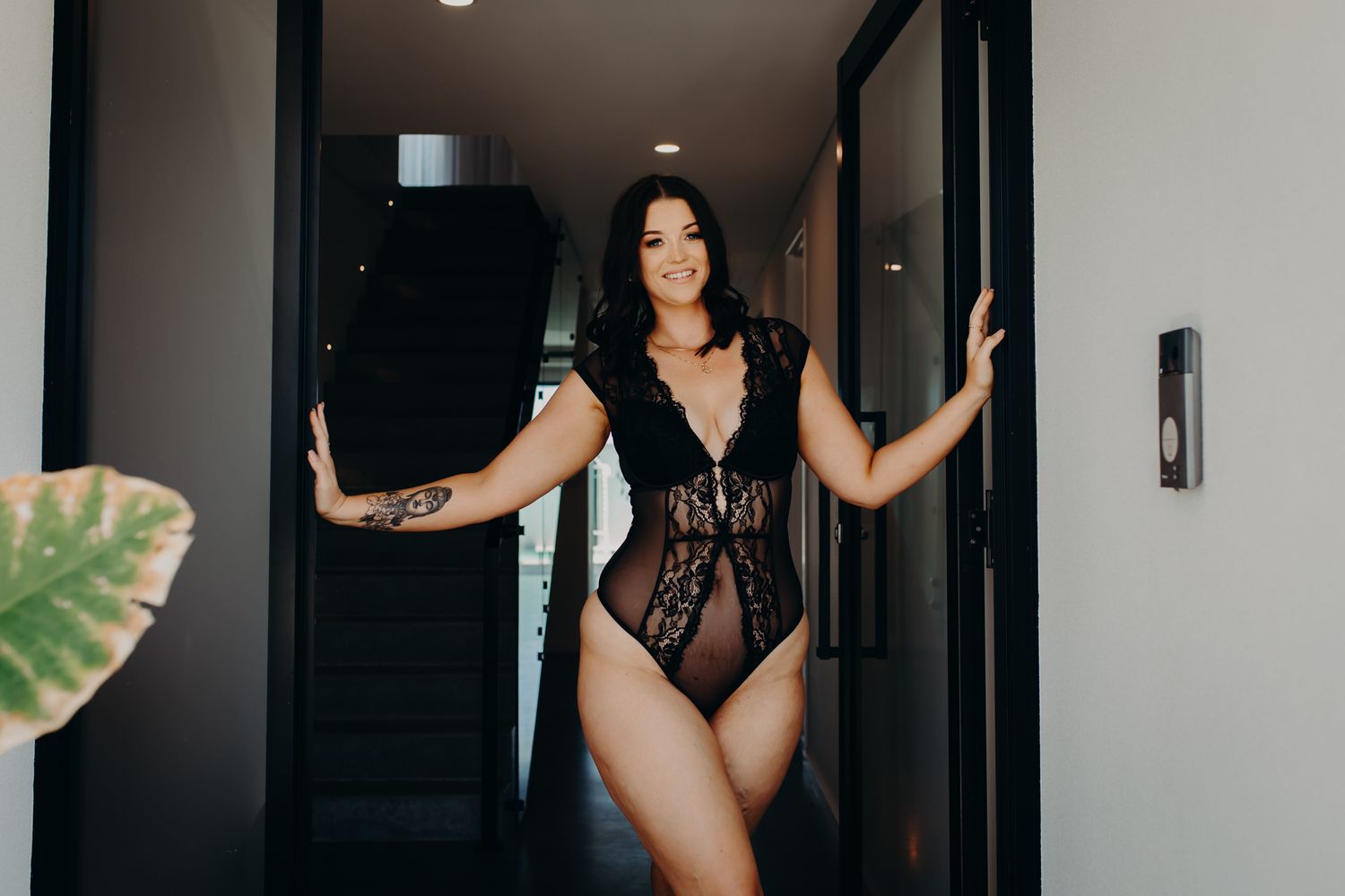 Person in black lace bodysuit posing in a hallway with houseplants.