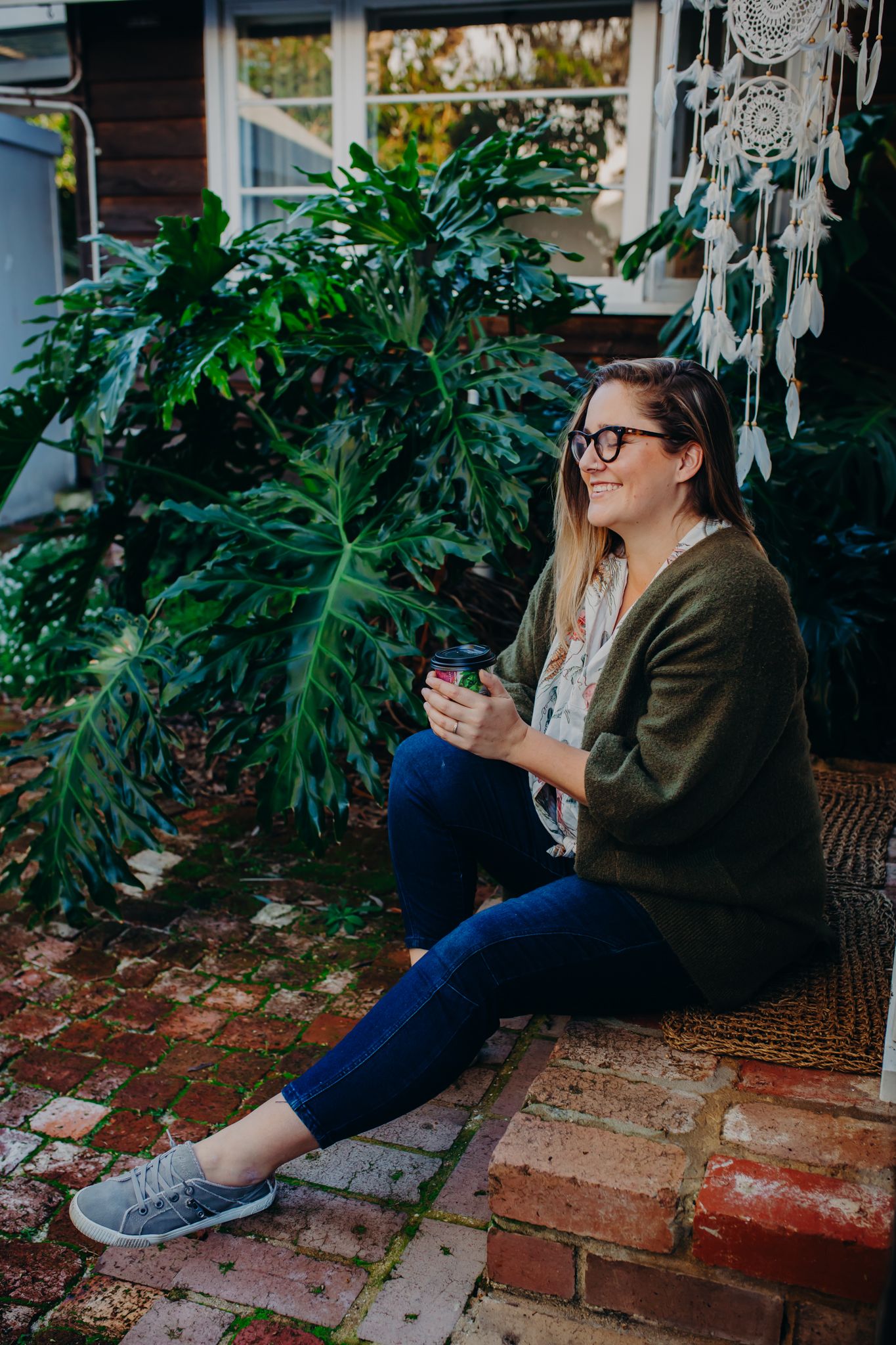 Person in cardigan and jeans sitting on brick steps near lush garden foliage.