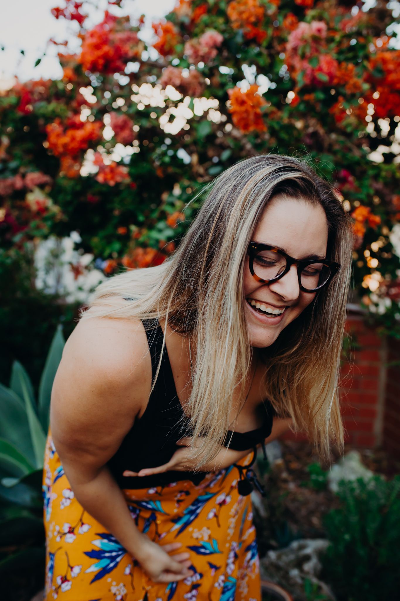 Person in glasses laughing against backdrop of vibrant flowering bushes.