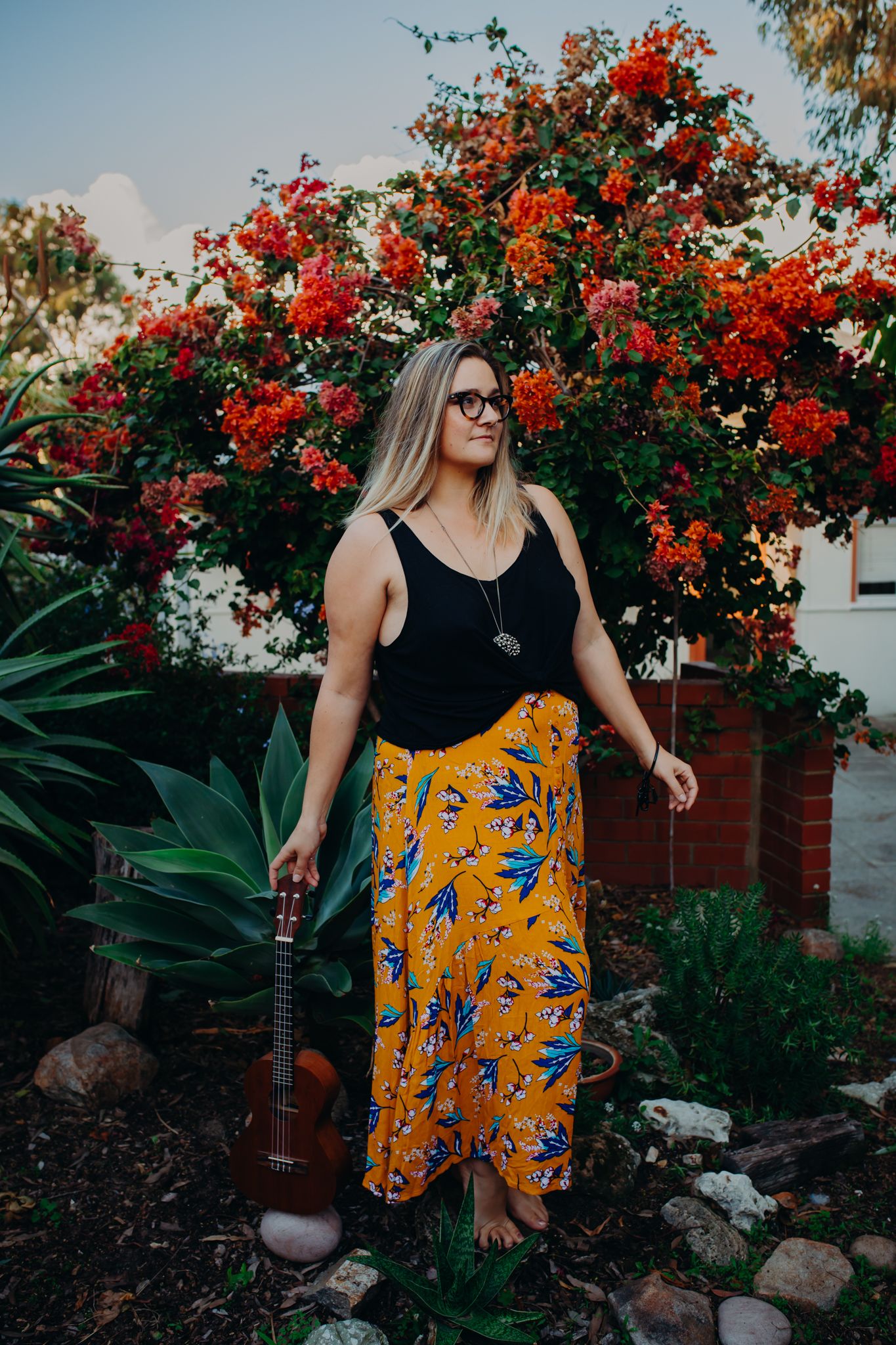 Person in yellow floral skirt and black tank top posing with ukulele in garden.
